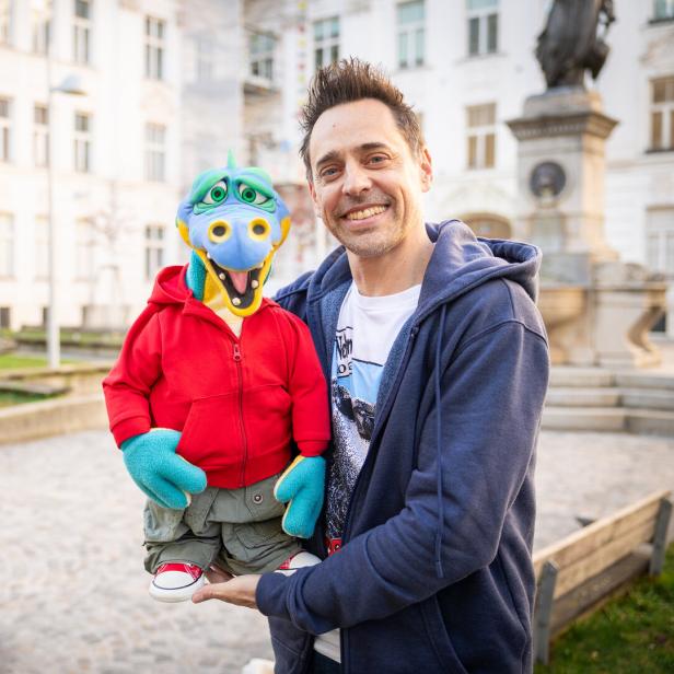 Ein Mann lächelt mit einer bunten Drachenpuppe im Arm vor einem Gebäude.