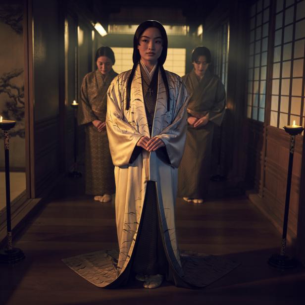 Eine Frau in einem Kimono steht in einem traditionellen japanischen Haus, flankiert von zwei weiteren Frauen.