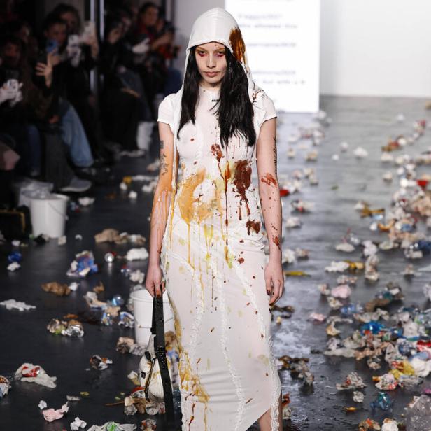 Ein Model mit Kapuze und einem mit Farbe bekleckerten Kleid auf einem Laufsteg voller Müll.