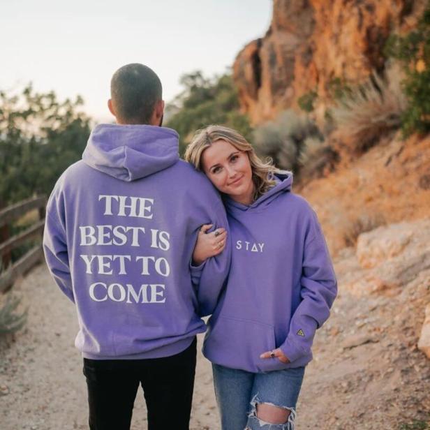 Ein Paar in lila Hoodies steht vor einer Felsformation; auf den Hoodies stehen die Worte „The best is yet to come“ und „Stay“.