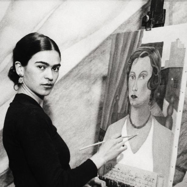 Frida Kahlo steht vor einer Staffelei und malt ein Porträt.