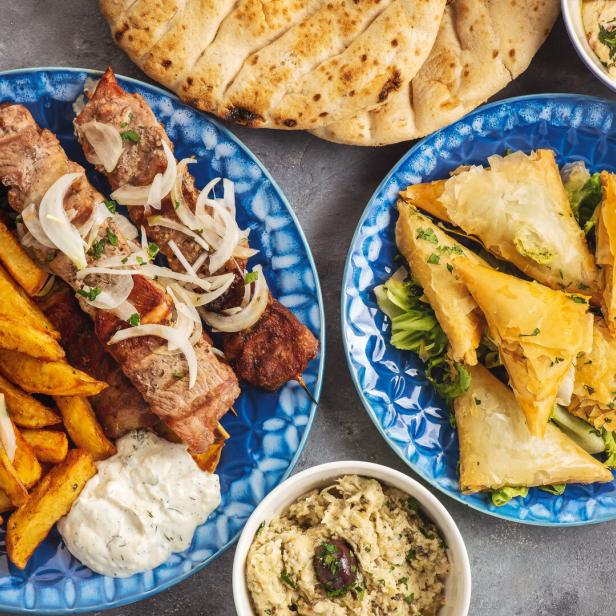 Zwei blaue Teller mit Souvlaki, Pommes, Salat, Blätterteiggebäck und Dips sowie Pitabrot.
