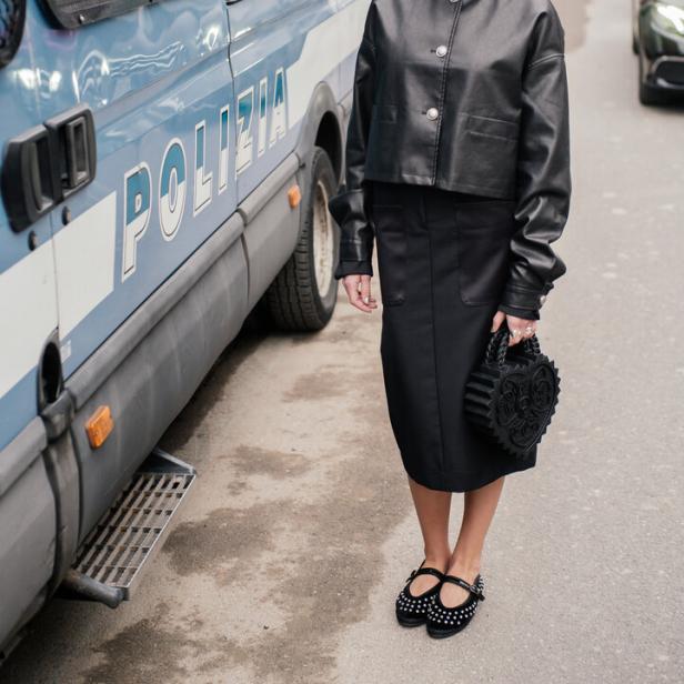Eine Frau in schwarzer Lederjacke und Rock steht neben einem Polizeiwagen.