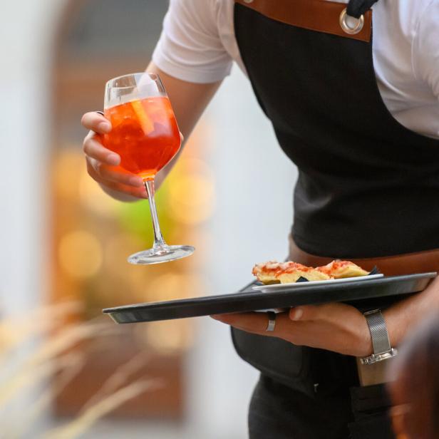 Ein Kellner serviert einen Aperol Spritz und kleine Pizzas im Schanigarten.