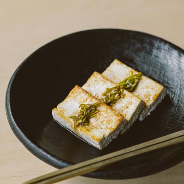 Gebackener Tofu mit grüner Garnitur auf einem schwarzen Teller.