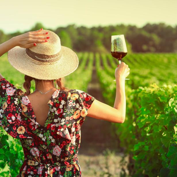 Frau im Sommerkleid und mit Sonnenhut hebt ein Weinglas mit Rotwein hoch, steht inmitten von Weinbergen.