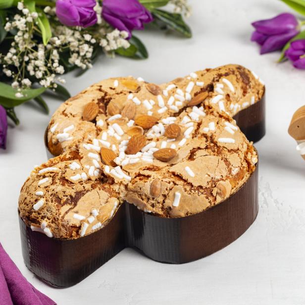 Eine Colomba Pasquale, ein traditioneller italienischer Osterkuchen, mit Mandeln und Zuckerstreuseln, neben einem Holzhasen und lila Tulpen.