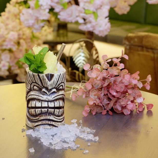 Ein Cocktail namens „Half Cuban“ in einem Tiki-Becher mit Eiswürfeln und rosa Blumen.