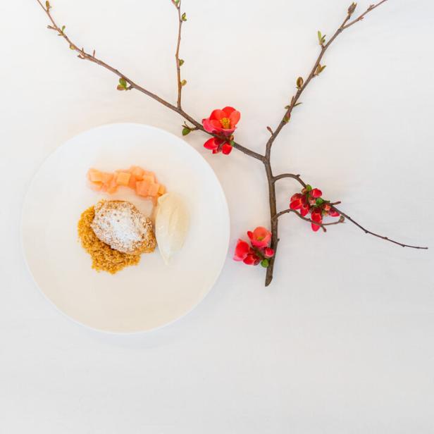 Ein Teller mit Dessert, bestehend aus Crumble, Eis und Rhabarber, dekoriert mit einem blühenden Zweig.