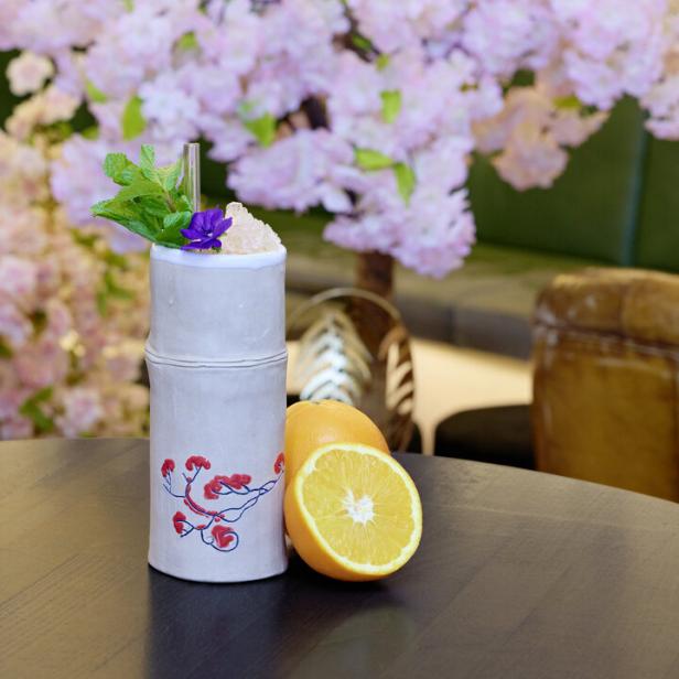 Ein Cocktail mit Minze und einer Blüte steht neben einer halben Orange vor einem blühenden Baum.