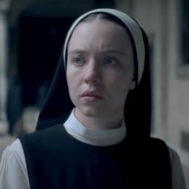 Eine junge Nonne mit ernstem Blick steht in einem Klostergang.