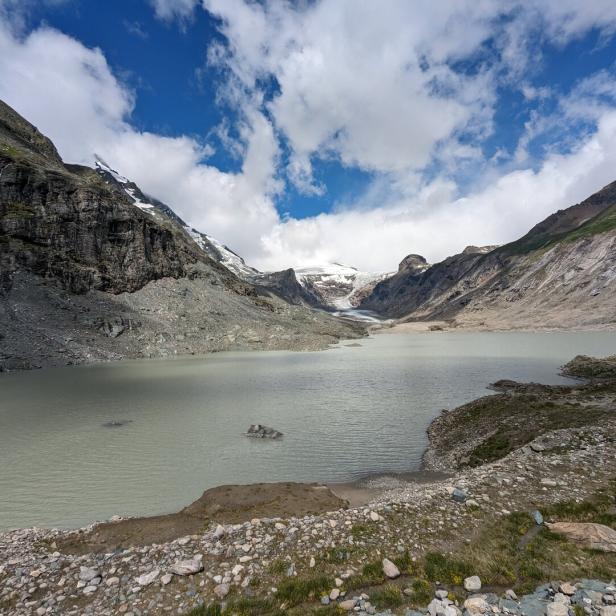 Ein Bergsee liegt eingebettet zwischen felsigen Hängen und einem Gletscher unter blauem Himmel.