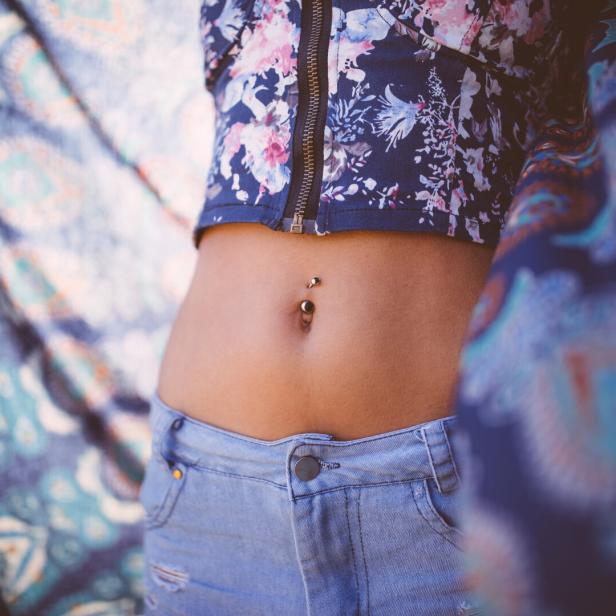Eine Frau mit einem Bauchnabelpiercing trägt ein kurzes, geblümtes Oberteil und Jeansshorts.