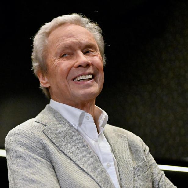 Ein lächelnder Mann mit grauem Haar trägt ein helles Jackett und eine Uhr am Handgelenk.