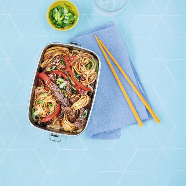 Honig-Chili-Rindfleisch-Nudeln in einer Lunchbox mit Stäbchen und einem Glas Wasser.