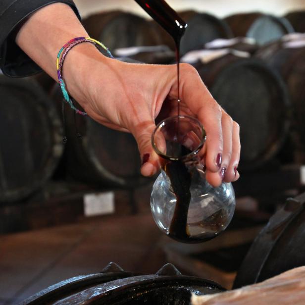 Hand mit Glasflasche und dunkler Flüssigkeit vor Fässern.