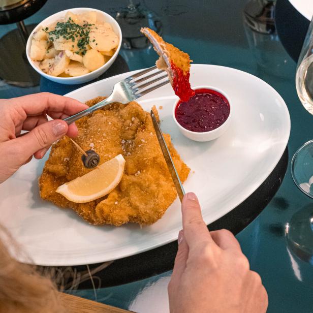 Eine Hand hält eine Gabel, worauf sich ein Schnitzelstück befindet. In der anderen ist ein Messer. Vor der Person liegt auf einem Teller ein schönes Wiener Schnitzel, garniert mit einer Zitronenspalte und einem Sardellenring.