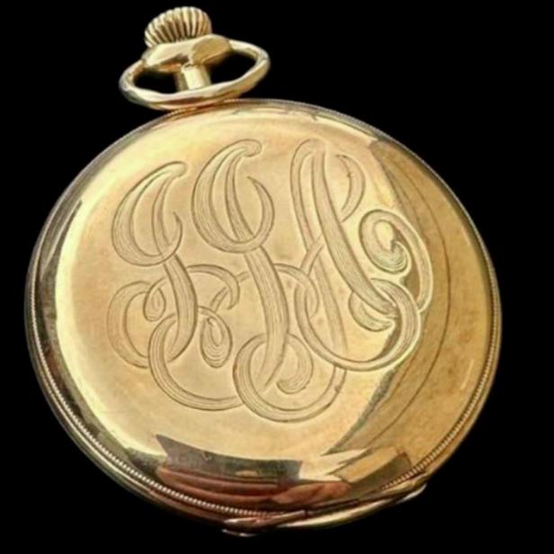 Eine goldene Taschenuhr mit eingraviertem Monogramm vor schwarzem Hintergrund.