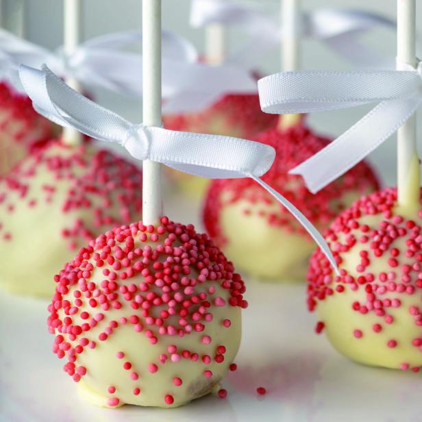 Mehrere Cake Pops mit weißer Glasur und rosa Streuseln, verziert mit weißen Schleifen.