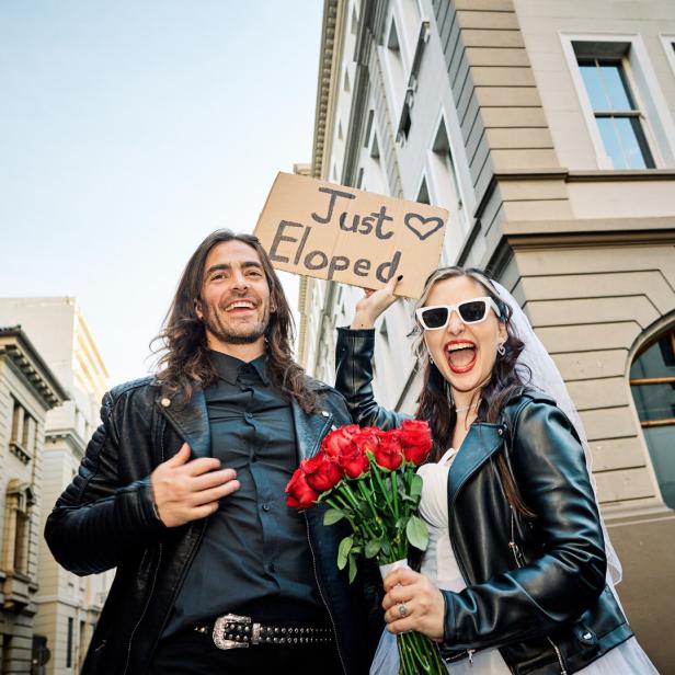 Ein frisch vermähltes Paar in Lederjacken hält ein Schild mit der Aufschrift „Just Eloped“.