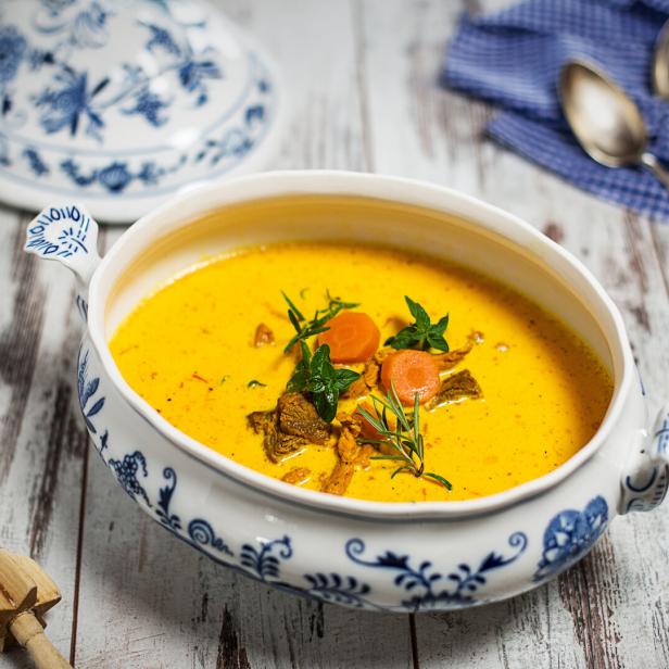 Eine Schüssel mit gelber Suppe, garniert mit Karotten und Kräutern, steht auf einem Holztisch.