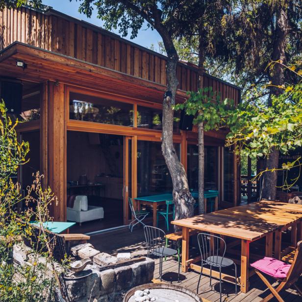 Ein Holzhaus mit Terrasse und Tischen im Freien, umgeben von Bäumen und Pflanzen.