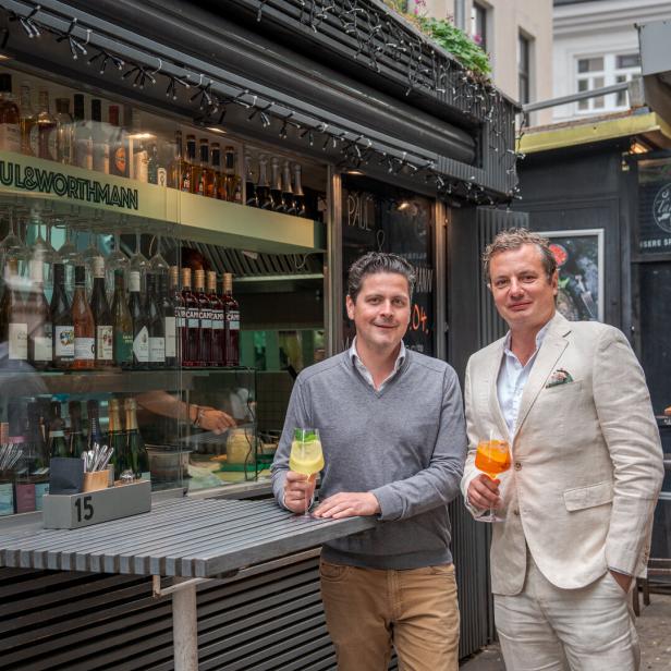 Zwei Männer stehen vor der Bar „Paul & Worthmann“ und halten Aperol Spritz in der Hand.