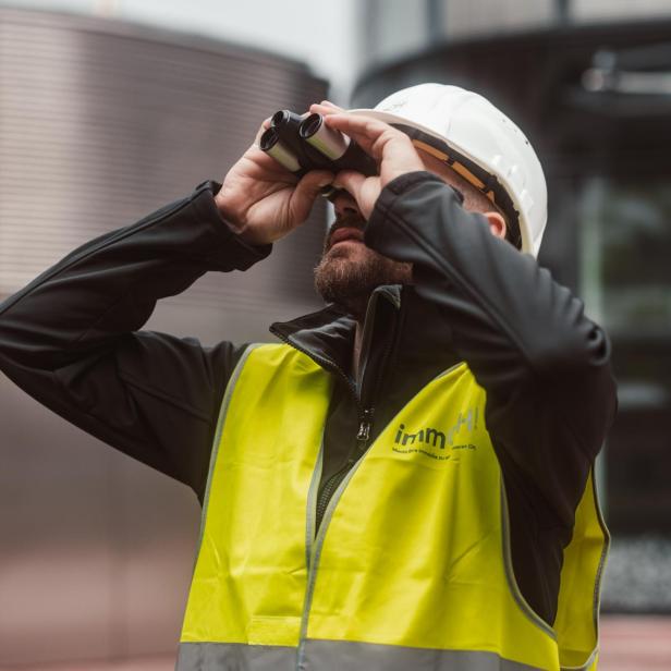 Bauarbeiter mit Helm und gelber Warnweste inspiziert etwas mit einem Fernglas
