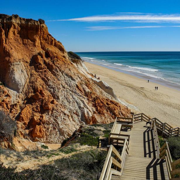 Eine Holztreppe führt zu einem Sandstrand mit roter Felswand unter blauem Himmel.