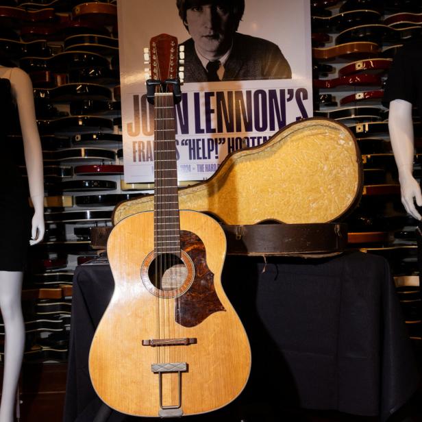 John Lennons Gitarre steht vor einem Poster von ihm und einem Gitarrenkoffer.