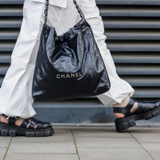 Eine Person mit weißen Hosen und schwarzen Sandalen trägt eine schwarze Chanel-Tasche.