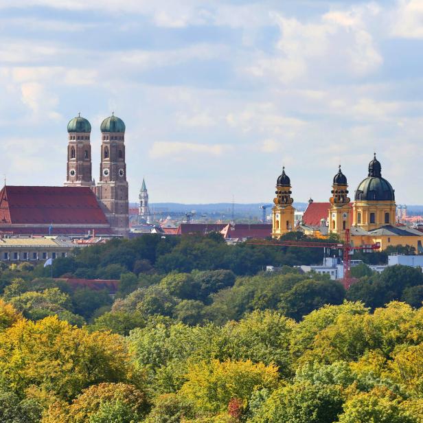 Blick über München mit Frauenkirche und anderen historischen Gebäuden hinter herbstlichen Bäumen.