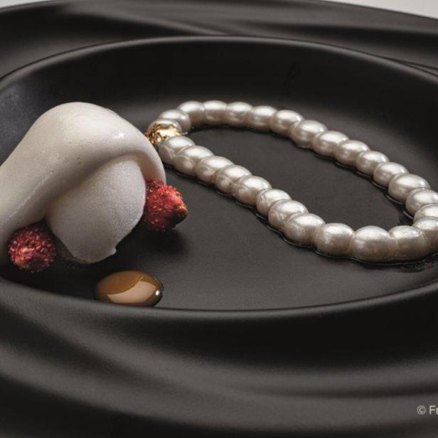 Ein Dessert mit Erdbeeren, Schaum und einer Perlenkette auf einem schwarzen Teller.