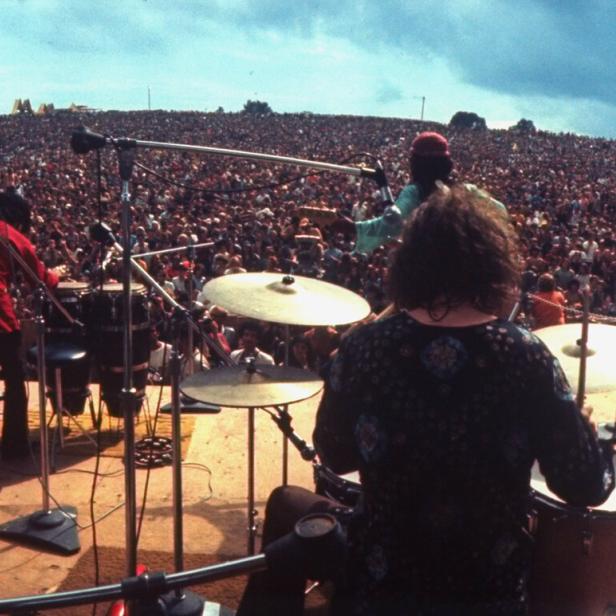Eine Band auf der Bühne beim Woodstock Festival 1969. Von der Bühne blicken die Musiker inmitten eine Menschenmasse.