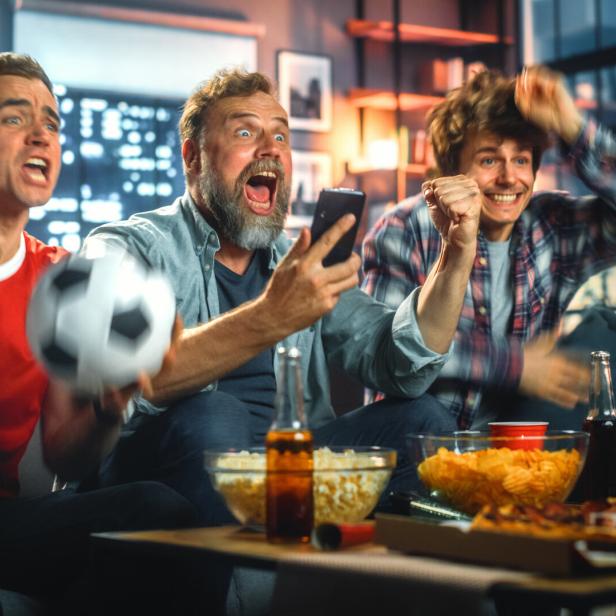 Drei Männer jubeln begeistert beim gemeinsamen Fußballschauen auf dem Sofa mit Snacks und Getränken.