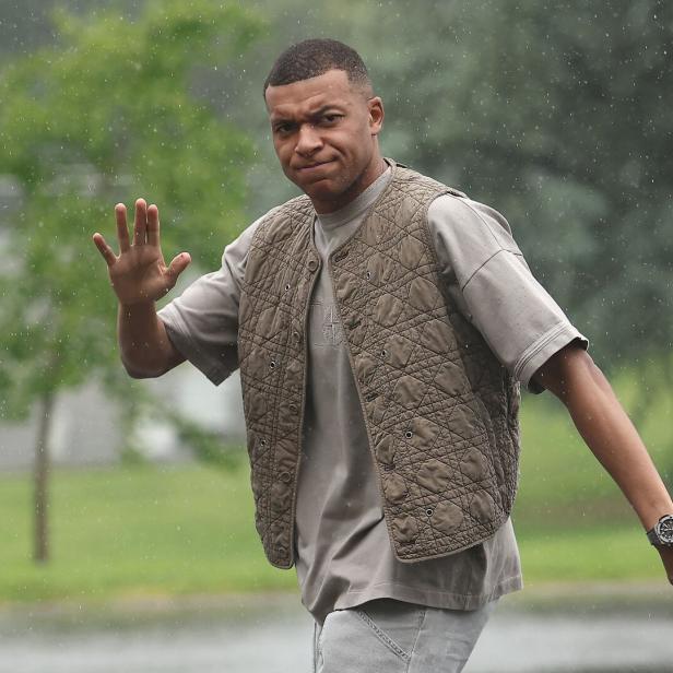 Fußballer Kylian Mbappé auf dem Weg zum Trainingslager in einer stylischen, beigen Weste.
