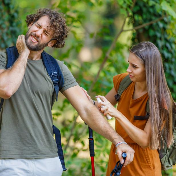 Eine Frau sprüht ihrem von Insektenstichen geplagten Wanderpartner Insektenspray auf den Arm.