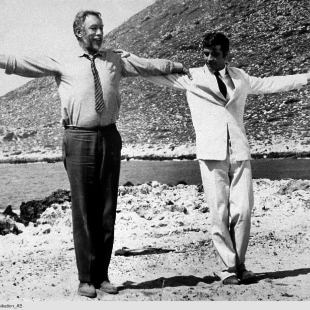 Zwei Männer, möglicherweise Darsteller des Films „Alexis Sorbas“, stehen am Strand mit ausgestreckten Armen.
