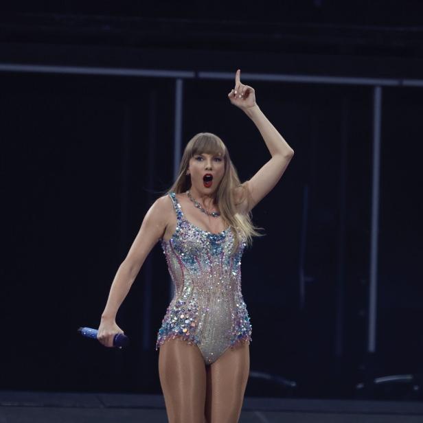 Taylor Swift steht auf der Bühne und zeigt mit dem Finger nach oben.
