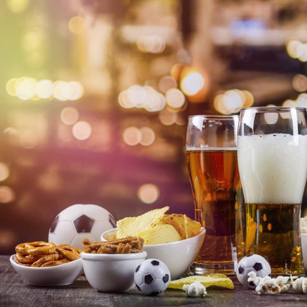 Zwei Gläser Bier, Snacks und Fußbälle auf einem Tisch vor verschwommenem Hintergrund.