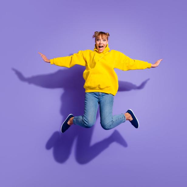 Junge Frau mit gelbem Hoodie springt fröhlich mit ausgebreiteten Armen vor lilafarbenem Hintergrund.