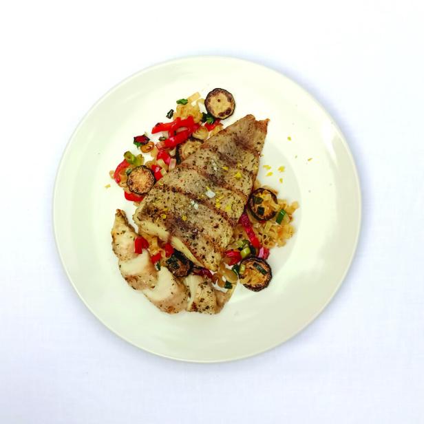 Ein Teller mit gegrilltem Fisch, Zucchini und Paprika auf einem weißen Tisch.