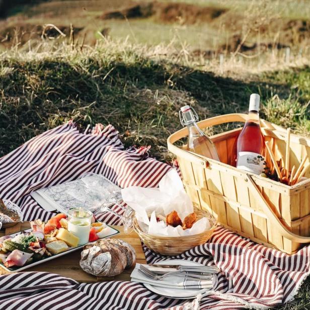 Ein Picknickkorb und Speisen auf einer rot-weiß gestreiften Decke im Grünen.