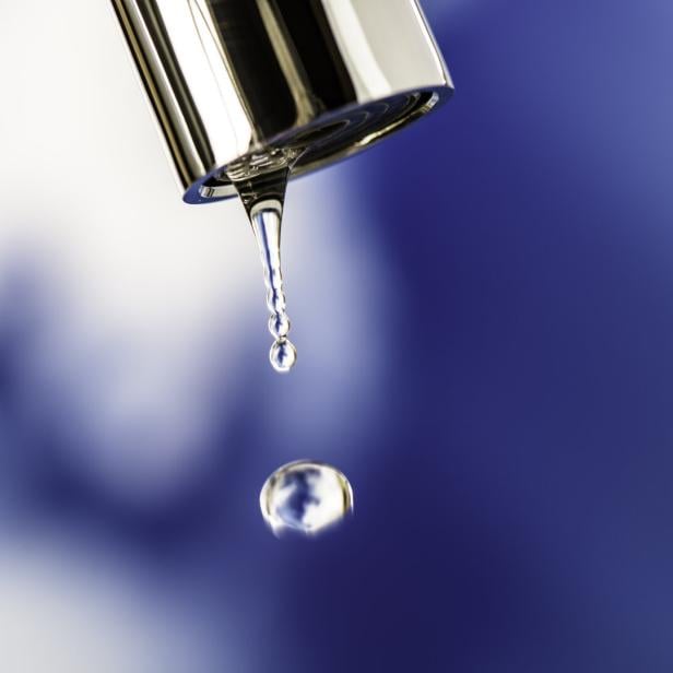 Ein Wassertropfen fällt aus einem silbernen Wasserhahn vor unscharfem blau-weißem Hintergrund.