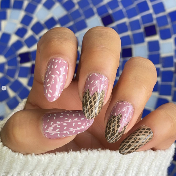 Eine Hand mit lackierten Nägeln im Eiscreme-Design vor blauem Mosaikhintergrund.