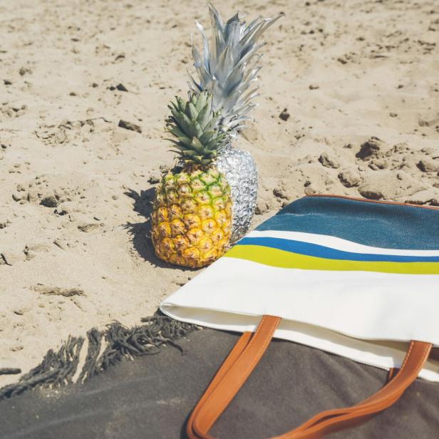 Eine Ananas und eine silberne Ananas-Skulptur liegen neben einer Strandtasche im Sand.