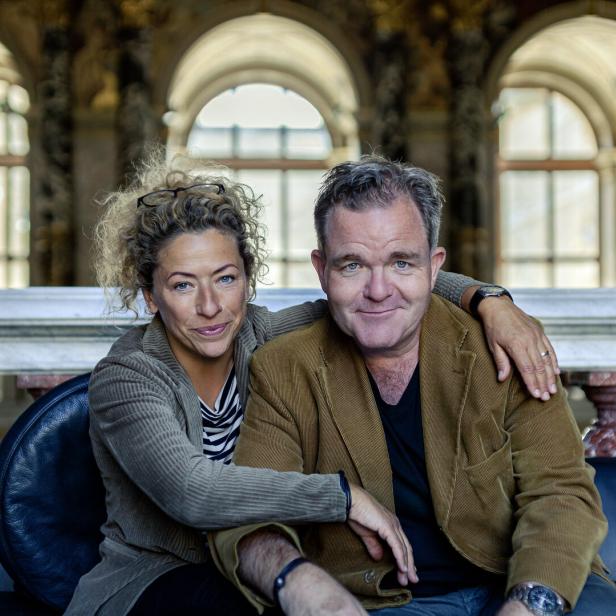 Carolin Pienkos und Cornelius Obonya sitzen auf einer Bank. Sie hat den Arm um ihn geschlungen, beide schauen sehr freundlich.