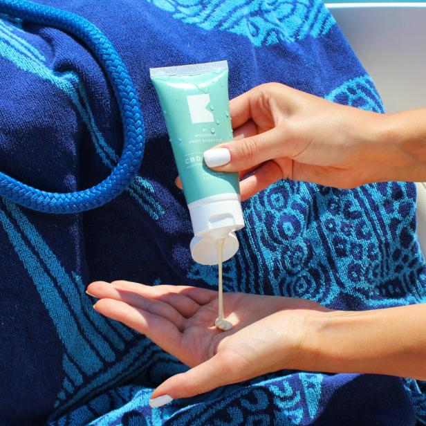 Eine Frau gießt CBD-Lotion in ihre Hand vor einem blauen Handtuch.