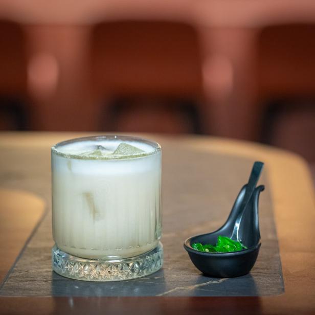 Ein Cocktail mit Eiswürfeln und eine Schale mit grünen Beilagen stehen auf einem Tisch.