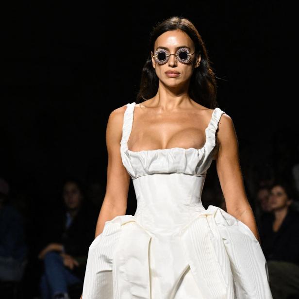 Irina Shayk trägt ein weißes Kleid und runde Sonnenbrille auf dem Laufsteg.